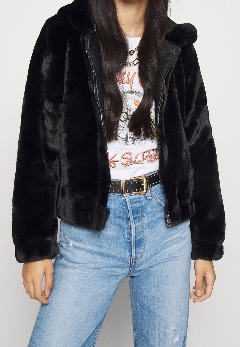 Personne portant une veste en fausse fourrure noire sur un t-shirt blanc graphique rentré dans un jean bleu clair taille haute avec une ceinture noire cloutée.