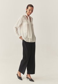 Biała jedwabna bluzka z kołnierzykiem na sznurek, z pionowymi plisami. Połączona z czarnymi szerokimi spodniami i czarnymi butami na obcasie z odkrytymi palcami.