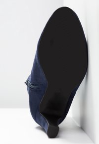 Bota de ante azul marino con cremallera lateral vista desde la suela, con un tacón bajo negro, sobre un fondo blanco.