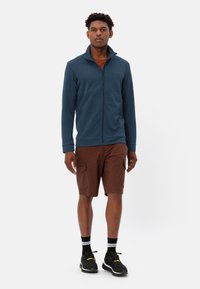 Giacca in pile blu con zip, pantaloni cargo marroni, scarpe da ginnastica nere con dettagli gialli, in piedi su uno sfondo bianco.