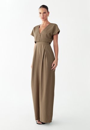 Femme debout portant une combinaison marron sur mesure avec manches courtes, encolure en V, boutons, jambes larges, et sandales noires à talons, sur fond blanc.