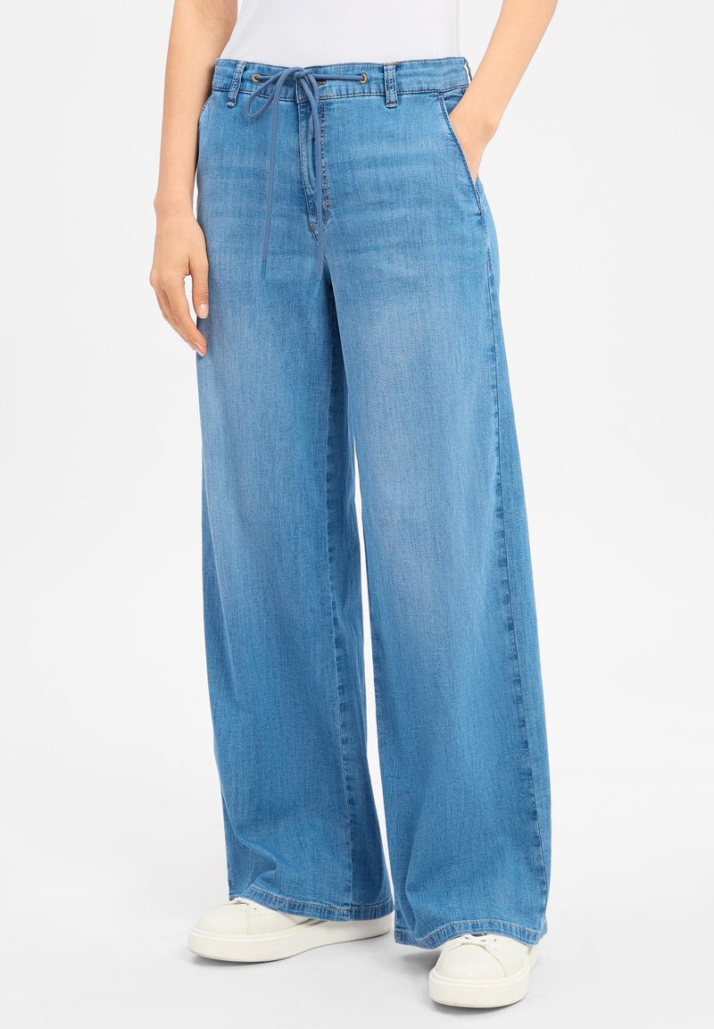 Hellblau weite Jeanshose mit Kordelzugbund, frontalen Taschen und einer weichen Textur. Getragen mit weißen Sneakers.
