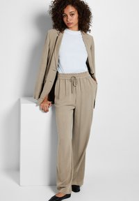 Beige, lichtgewicht pak met een gepaste blazer en losse, elastische broek. Witte T-shirt met ronde hals eronder. Zwarte ballerina's.