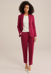 Fuchsia kostym med skräddarsydd kavaj och smala byxor, kombinerat med en vit kofta. Glansiga vita klackar fulländar looken.