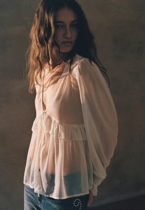 Junge Frau mit langen, welligen Haaren, die eine lockere, transparente weiße Bluse und blaue Jeans trägt, vor einem gedämpften, neutralen Hintergrund steht.