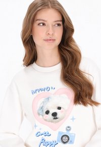 Weißes Sweatshirt mit einer großen Grafik eines Welpenkopfes mit einer blauen Schleife, umgeben von rosa Herz- und Kameraillustrationen.