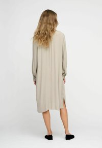 Light beige long-sleeve dress with a loose fit, side slits, and soft, flowing fabric. Worn with black slip-on shoes.