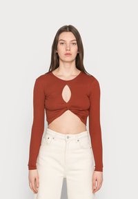 Top cropped de cor ferrugem com nervuras e mangas longas, apresentando um recorte em forma de chave no centro e detalhes torcidos na barra. Combinado com calças brancas.
