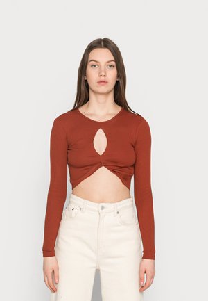 Top cropped de cor ferrugem com nervuras e mangas longas, apresentando um recorte em forma de chave no centro e detalhes torcidos na barra. Combinado com calças brancas.