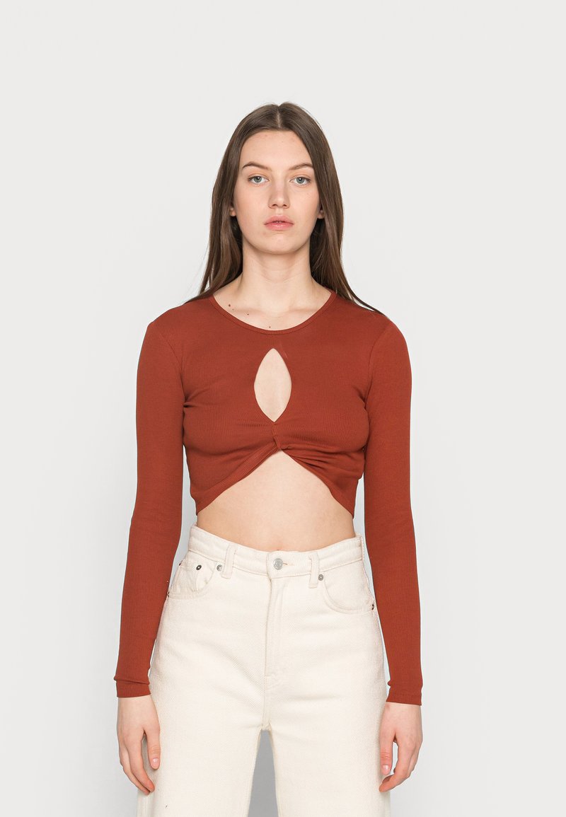 Top cropped de cor ferrugem com nervuras e mangas longas, apresentando um recorte em forma de chave no centro e detalhes torcidos na barra. Combinado com calças brancas.