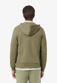 Felpa verde oliva con finitura testurizzata, cappuccio con cordino regolabile e polsini a coste. Sovrapposta a una camicia a righe, vista da dietro.