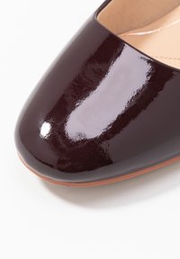 Chaussure plate en cuir verni bordeaux brillant avec un bout arrondi et une fine semelle beige. Pas d'ornements ni de motifs visibles.