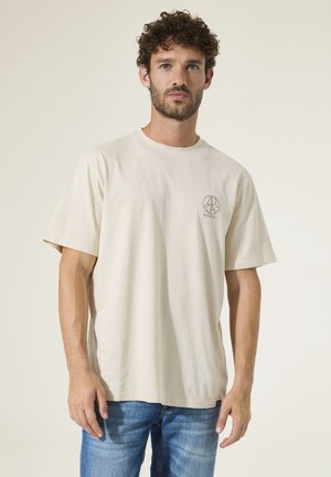Man met krullend haar, die een beige T-shirt met korte mouwen draagt met een klein logo op de borst en blauwe spijkerbroek, staand tegen een effen lichte achtergrond.