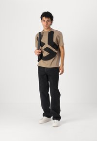 Brown cotton t-shirt with large black graphic design, paired with black denim pants and white sneakers. Model holds a black bag.
