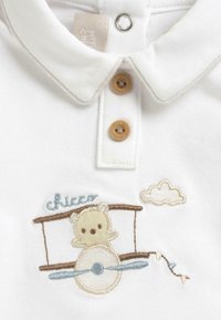 Polo bébé blanc avec deux boutons beige, représentant un ours en peluche brodé pilotant un avion marron et bleu avec un petit nuage au-dessus.