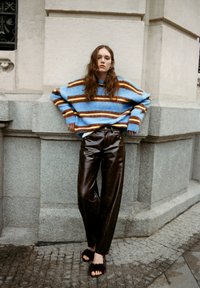 Pulover mare tricotat în dungi albastre și maronii, pantaloni din piele neagră și sandale pufoase închise, pozat contra unui zid de piatră texturat.