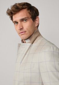 Mężczyzna z jasnobrązowymi włosami nosi beżową marynarkę w kratę z uniesionym kołnierzem, lekko patrząc w lewo na jednolity, jasny tle.