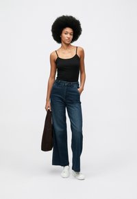 Kvinne med afro-frisyre iført svart topp med stropper, vide blå jeans, hvite joggesko, og holder en brun veske foran en hvit bakgrunn.