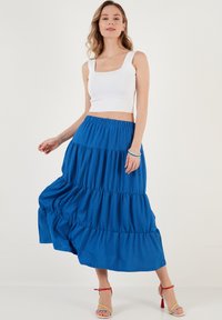 Kék, réteges maxi szoknya, rugalmas derékkal, fehér, négyzet alakú kivágású crop toppal párosítva. A modellen színes, pántos magassarkúk vannak.