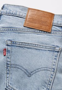 Lyseblå denimjeans med et brunt lærmerke med preget tekst, rød Levi's-tab, og kontrastsøm på baklommene.