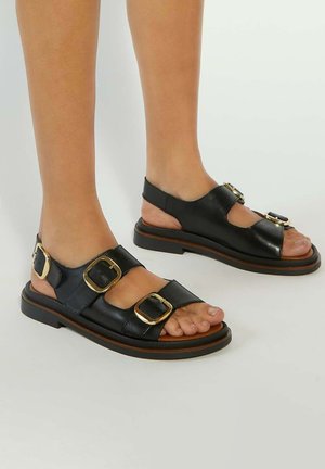 Black leather sandals with three gold buckles, open-toe design, and a low brown rubber sole; textured upper and adjustable back strap.