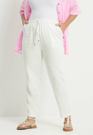 Kvinde iført hvide linnedbukser med snøre, hvid top, lyserød skjorte med knapper og beige sandaler med stropper, stående med hænderne i lommerne.