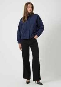 Marinblå kviltad jacka med hög krage, elastiska ärmmuddar och två framfickor. Matchad med svarta vida byxor och spetsiga svarta klackar.