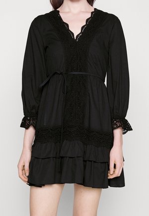 Woman wearing a black long-sleeve dress with lace trim on neckline, cuffs, and waist, featuring a tiered ruffle skirt and a waist tie.