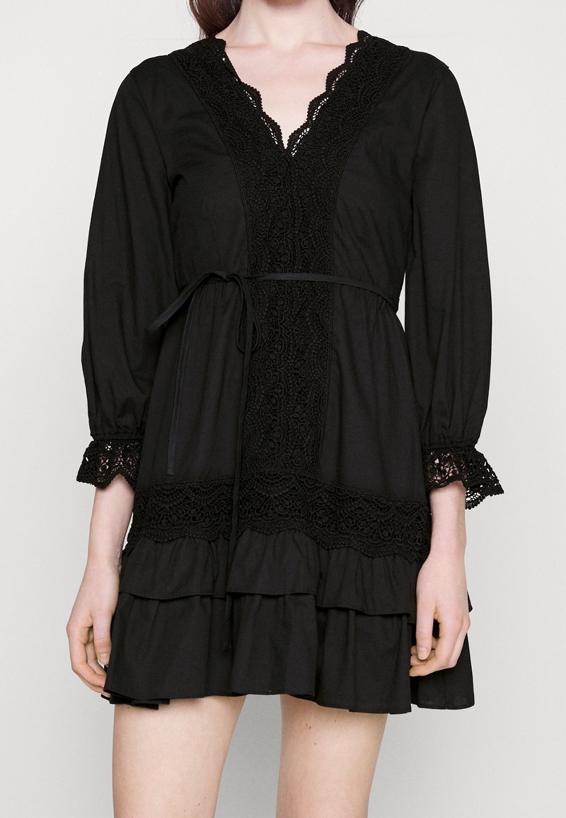 Femme portant une robe noire à manches longues avec bordure en dentelle au niveau de l'encolure, des poignets et de la taille, présentant une jupe à volants superposés et une ceinture à la taille.