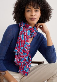 Röd och blå mönstrad scarf i lätt tyg med fransade kanter, draperad runt halsen, som kompletterar en strukturerad marinblå tröja.