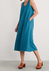 Robe midi sans manches en turquoise avec une coupe décontractée et des poches latérales, portée avec des sabots en bois brun sur un fond neutre.