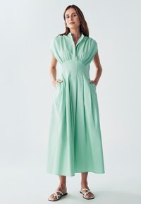 Mätovo zelené šaty s krátkym rukávom, s gombíkovým golierom, elastickým pásom, bočnými vreckami a splývavou dĺžkou maxi. Hladká, ľahká tkanina.