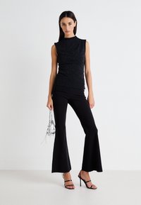 Top negru fără mâneci cu guler înalt și strălucire subtilă, asortat cu pantaloni negri evazați. Accesoriile includ o mică poșetă argintie.