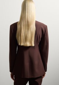Blazer marrón ajustado con hombros estructurados y una textura suave, combinado con pantalones a juego. Larga melena rubia recta cae por la espalda.
