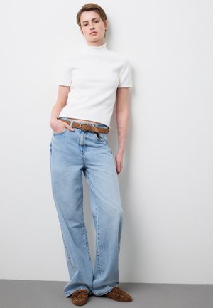 Joven con cabello corto apoyado en la pared, lleva un suéter de cuello alto blanco de manga corta, jeans de pierna ancha azul claro, cinturón marrón y mocasines.