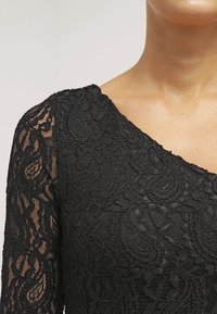 Haut en dentelle noire à manches longues, présentant des motifs cachemire complexes et un large décolleté. La texture transparente met en valeur le design détaillé.