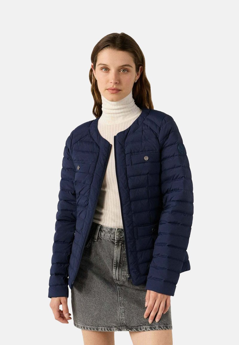 Giacca imbottita trapuntata blu navy con collo tondo, chiusura con bottoni a pressione e due tasche sul petto. Abbinata a un dolcevita bianco a coste e una gonna di denim.