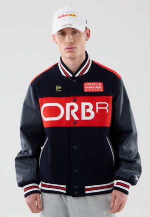 Mladý muž v čiernej a červenej bunde Oracle Red Bull Racing a bielej šiltovke Oracle Red Bull Racing stojí pred jednofarebným pozadím.