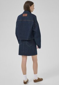 Ensemble en denim comprenant une veste et une jupe de couleur bleu foncé, avec une forme carrée, un design oversize, une étiquette en cuir au dos et des poches arrière.