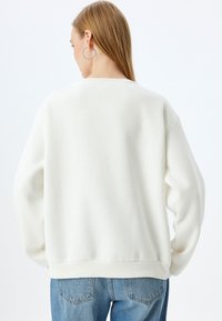 Sudadera blanca de gran tamaño con puños y dobladillo de canalé, que presenta una textura suave. Combinada con jeans de un azul claro, vista desde atrás.