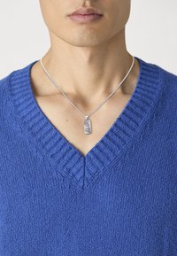 Pulover bleu tricotat cu un decolteu în formă de V, având manșete cu nervuri. Colier din argint cu un pandantiv dreptunghiular texturat.
