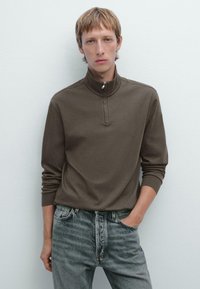 Brauner Zip-Kragen-Pullover aus weichem Material mit langen Ärmeln, kombiniert mit ausgebleichten hellgrauen Jeans. Einfaches Design, ohne auffällige Muster.