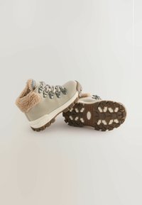 Beige vinterstøvler med pelsfor, metalsnørehuller og tykke brune gummisåler med dybe mønstre for godt greb, placeret side om side.