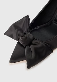 Ballerine nere in satin con punta a punta e un grande dettaglio a fiocco sul dorso del piede, caratterizzate da una texture liscia e foderatura interna, design minimalista.