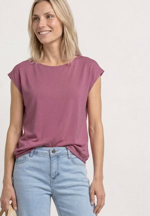 Uśmiechnięta kobieta z blond włosami do ramion, ubrana w mauve'owy top z krótkim rękawem i jasnoniebieskie jeansy, trzymająca plecioną torebkę w lewej ręce.