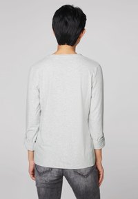 Světle šedá dlouhá rukávová košile s ohrnutými rukávy a kulatým výstřihem. Měkká, texturovaná látka. Zadní strana má jednoduchý, čistý design.