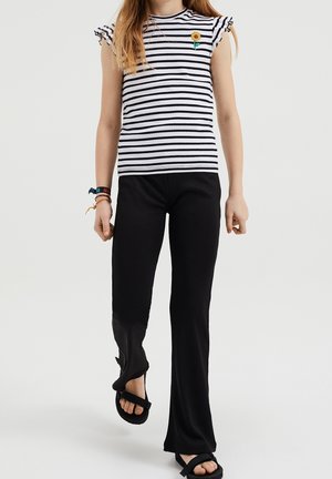 Jeune personne portant des sandales noires, un pantalon côtelé noir avec une fente à l'avant, et une chemise à rayures blanches et noires avec un patch tournesol sur la poitrine.