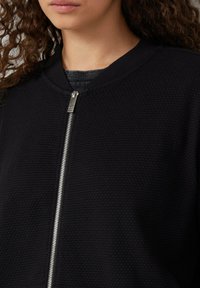 Veste bomber noire en tissu texturé avec fermeture éclair à l'avant, col côtelé et détails de fermeture éclair lisse en argent.