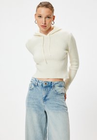 Kürzer geschnittene, gerippte, cremefarbene Kapuzenjacke mit Kordelzug. Kombiniert mit hoch geschnittenen, verblassten blauen Jeans mit Knopfverschluss und Taschen.
