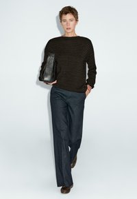 Persona che indossa un maglione a strisce nere e marroni, pantaloni larghi scuri, scarpe marroni e tiene una borsa in pelle nera su uno sfondo bianco uniforme.